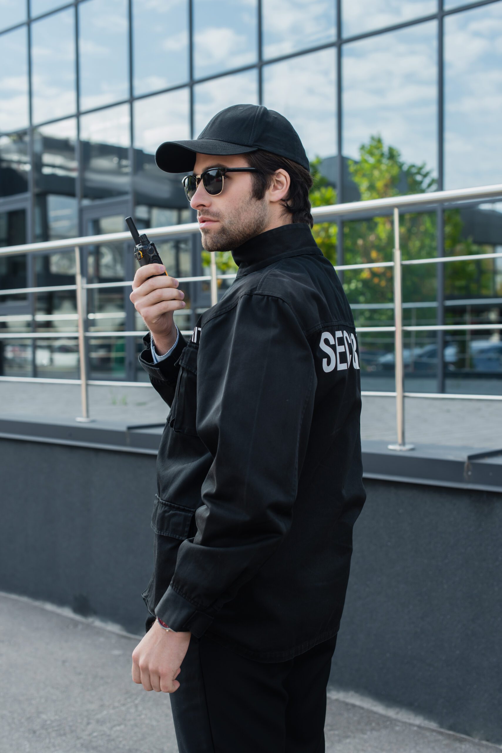 young security man in uniform, cap and sunglasses talking on walkie-talkie near building young security man in uniform, cap and sunglasses talking on walkie-talkie near building